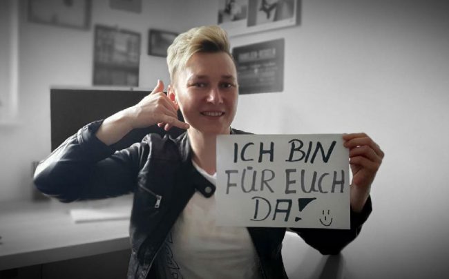 Mitarbeiterin mit Papierschild Ich bin für euch da und die Finger zum Telefonhörer gebildet